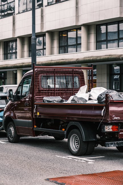 A dark red flatbed truck parked on a city street in front of a modern office building with large glass windows and white architectural panels. The truck's open cargo area is filled with various types of waste, including black and white plastic bags, some of which appear crumpled or partially opened, and miscellaneous debris. The vehicle features a metal safety grille behind the cab and small wheels with silver hubcaps. The surroundings include a lamppost to the left and other parked vehicles in the background. The scene captures an urban environment consistent with private waste collection activities, illustrating a typical scene of rubbish removal by a professional service such as Rubbish Removal Haringey, highlighting an alternative or on-site clearance option for bulky waste disposal in the Haringey area.