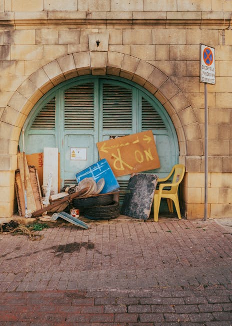 A collection of discarded waste is piled in front of a large, rounded, teal-colored metal door set into a sandstone building wall. The rubbish includes wooden planks, a flipped blue plastic container, a faded yellow sign with partially visible writing, and a black tire placed on the ground. A yellow plastic chair leans against a black object among the debris, which covers part of a paved sidewalk. To the right of the waste, a metal pole with a no-parking sign is mounted on the wall. The scene appears to be an outdoor area adjacent to a historic or older building, with natural daylight illuminating the scene. This setting is indicative of private or independent waste disposal, often managed by services like Rubbish Removal Haringey, rather than municipal rubbish collection.