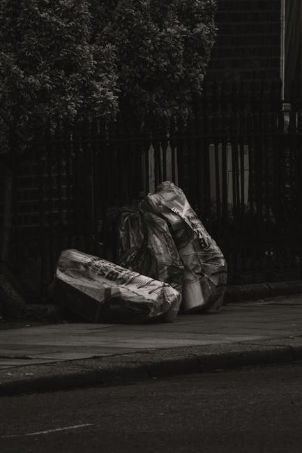 Three large black garbage bags filled with waste are placed on a paved sidewalk near a black metal fence and a dense hedge with leafy foliage. The bags appear to be made of plastic, with some showing signs of being tightly knotted or gathered at the tops, suggesting they are ready for collection. The scene is set outdoors, possibly at the edge of a residential or commercial property, with a dark background behind the fence and minimal natural light, giving the image a subdued, almost monochrome appearance. The bags are positioned in the foreground, with the hedge and the fence providing a contrasting backdrop, indicating an outdoor rubbish collection point that could be managed by a private waste removal service such as Rubbish Removal Haringey. Overall, the image portrays a typical on-street waste disposal scene aligned with non-LEA rubbish clearance methods, emphasizing the importance of proper waste management and private collection options in urban settings.