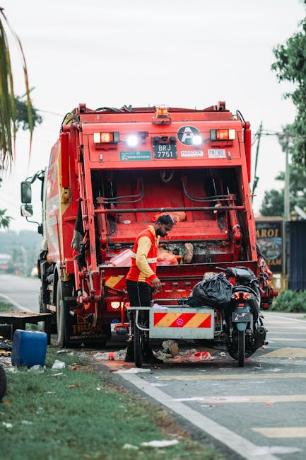 A worker in a yellow and red uniform is operating a large red rubbish collection truck on a roadside in a suburban area, with green trees and utility poles in the background. The worker is standing at the rear of the truck, which is equipped with mechanical lifting mechanisms for waste collection. The truck's rear panel is open, revealing internal components and a metal hopper used for waste intake. A black garbage bag is placed in front of the worker on a small platform, and scattered litter is visible around the base of the truck, including trash and debris on the pavement. The scene appears to be part of an outdoor waste management or rubbish removal operation, possibly involving private or alternative waste disposal services, as indicated by the presence of the specialized vehicle and the worker's activity. The lighting is natural with an overcast sky, and the environment suggests an organized effort to clear rubbish from the area, aligning with services offered by companies like Rubbish Removal Haringey.