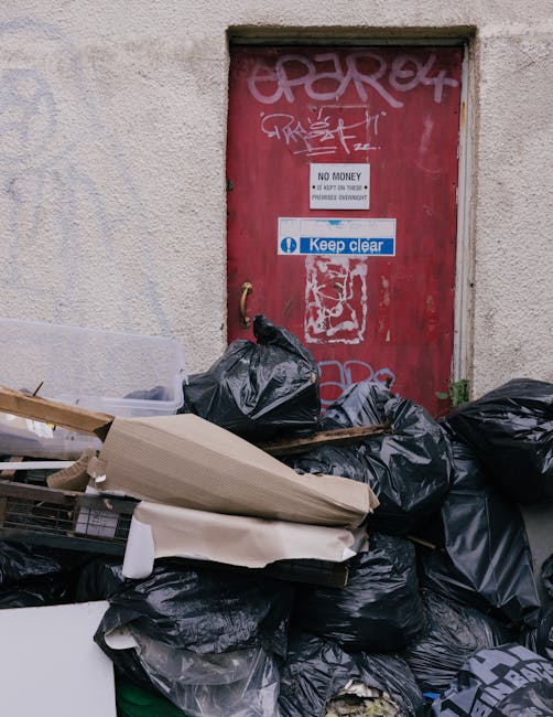Three large black garbage bags filled with waste are placed on a paved sidewalk near a black metal fence and a dense hedge with leafy foliage. The bags appear to be made of plastic, with some showing signs of being tightly knotted or gathered at the tops, suggesting they are ready for collection. The scene is set outdoors, possibly at the edge of a residential or commercial property, with a dark background behind the fence and minimal natural light, giving the image a subdued, almost monochrome appearance. The bags are positioned in the foreground, with the hedge and the fence providing a contrasting backdrop, indicating an outdoor rubbish collection point that could be managed by a private waste removal service such as Rubbish Removal Haringey. Overall, the image portrays a typical on-street waste disposal scene aligned with non-LEA rubbish clearance methods, emphasizing the importance of proper waste management and private collection options in urban settings.
