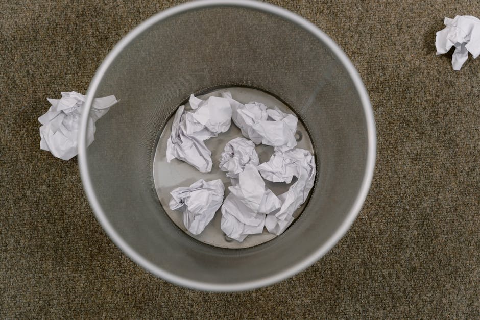 An overhead view of a round, metallic waste bin positioned on a textured brown carpeted floor. Inside the bin are several crumpled sheets of white paper, some of which are also scattered on the floor around the bin. The interior of the bin appears smooth with a matte finish, and the crumpled paper varies in size and shape, indicating discarded office waste. The scene suggests an indoor environment, possibly an office or work area, where waste has been temporarily collected for collection or disposal. The lighting is neutral, without shadows or reflections, emphasizing a straightforward, unobstructed view of the trash items and the bin. This visual context supports the process of private or alternative waste handling services offered by Rubbish Removal Haringey, aligning with the theme of removing office or general rubbish effectively without using standard council collection methods.