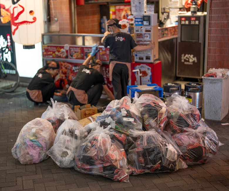 Multiple large transparent plastic bags filled with various discarded clothing items and textiles are stacked on a paved sidewalk area outside a commercial storefront. The bags are casual, crumpled, and loosely tied, revealing a mix of colourful fabrics and materials inside. Behind the bags, three workers wearing black uniforms with beige aprons and gloves are engaged in a task involving a red and silver automated machine or collection point, possibly for sorting or processing waste. The background includes a small food outlet with illuminated signage, a vending machine, and additional waste containers located against red-tiled walls. The environment appears to be an urban street scene, illuminated by artificial lighting, with the scene suggesting an instance of on-site waste collection or equipment for private rubbish handling, relevant to independent rubbish removal services like those offered by Rubbish Removal Haringey, focusing on waste management within commercial or retail premises.