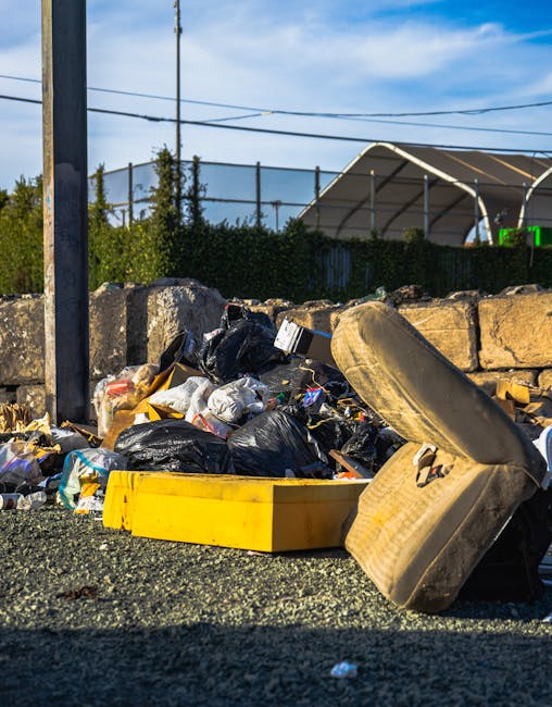 A pile of mixed waste materials and rubbish bags is accumulated on a gravel surface next to a short stone wall, with a large, beige, fabric-covered armchair lying on its side among the debris. The rubbish includes black trash bags, cardboard boxes, plastic containers, and loose household waste, all scattered in a disorganized manner. In the background, a metal utility pole with electrical wires extends into a partly cloudy sky, and behind the stone wall, there is a green hedge and a large, curved, transparent-roofed structure, possibly a storage or sports facility, partly obscured by the foliage. The scene appears to be an outdoor area for rubbish disposal or collection, illustrating issues with unmanaged waste accumulation that a private rubbish removal service such as Rubbish Removal Haringey might aim to address as part of alternative waste handling solutions in the borough.