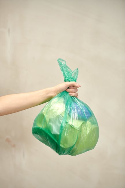 A large accumulation of black plastic rubbish bags piled against a tiled wall in an outdoor urban setting, with some partially torn open, revealing discarded plastic bottles and other trash inside. The bags are stacked haphazardly, occupying most of the foreground, and their shiny, crumpled surfaces reflect ambient light. Behind the pile, the wall features graffiti with the word 'XEND' spray-painted in red. To the left, a section of a metallic pole or pipe is visible, and the sky above shows dark, stormy clouds, indicating deteriorating weather conditions. The scene suggests a private or alternative waste disposal situation, emphasizing issues related to rubbish collection delays or mishandling. The environment appears to be an external area adjacent to a building, possibly a back alley or service zone, where litter has accumulated due to ineffective rubbish removal services, highlighting the importance of professional rubbish clearance such as that provided by Rubbish Removal Haringey.