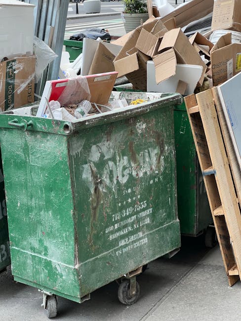 A large green waste disposal container with visible wear and rust on its surface, positioned on a sidewalk. The container is filled with various discarded materials, including flattened cardboard boxes, some with printed labels, and other packaging debris. The cardboard boxes are of different sizes, some open and leaning outward, revealing a mix of plain and printed exteriors in brown and white tones. To the right of the container, there is a wooden pallet stacked with more cardboard, leaning against the side of the container. In the background, blurred elements of an urban environment are visible, including a storefront window, a potted plant, and a parked vehicle on the street. The scene suggests an area undergoing private rubbish collection or on-site clearance, possibly handled by a professional waste removal service such as Rubbish Removal Haringey, with typical materials intended for disposal or recycling.