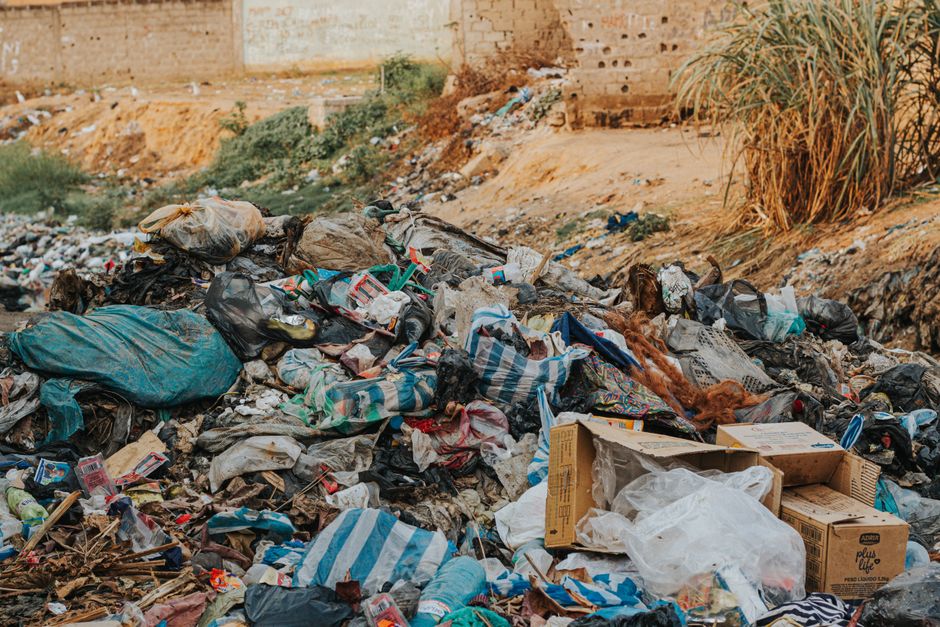 The image depicts a large amount of mixed household and construction waste discarded in an open outdoor area, characterized by a combination of plastic bags, cardboard boxes, torn fabric, and various loose debris. Many black and blue rubbish bags are visibly torn or partially collapsed, revealing their contents such as packaging, paper, and miscellaneous waste. The cardboard boxes are flattened or partially broken open, some with clear labels or printed text, and are situated amidst scattered plastic packaging, textiles, and crushed items. The surface beneath the rubbish appears uneven and dirt-colored, with patches of bare earth, small rocks, and sparse vegetation. In the background, a weathered brick wall and a few tall, dry grasses growing along the edge of the scene are visible, suggesting an urban or semi-urban environment. The lighting is natural, likely from daylight, illuminating the refuse and casting subtle shadows. This scene is indicative of unmanaged waste accumulation, often associated with illegal dumping or neglected disposal, which could be relevant to private rubbish removal services, such as those provided by Rubbish Removal Haringey, to facilitate off-site clearance or alternative waste handling methods.