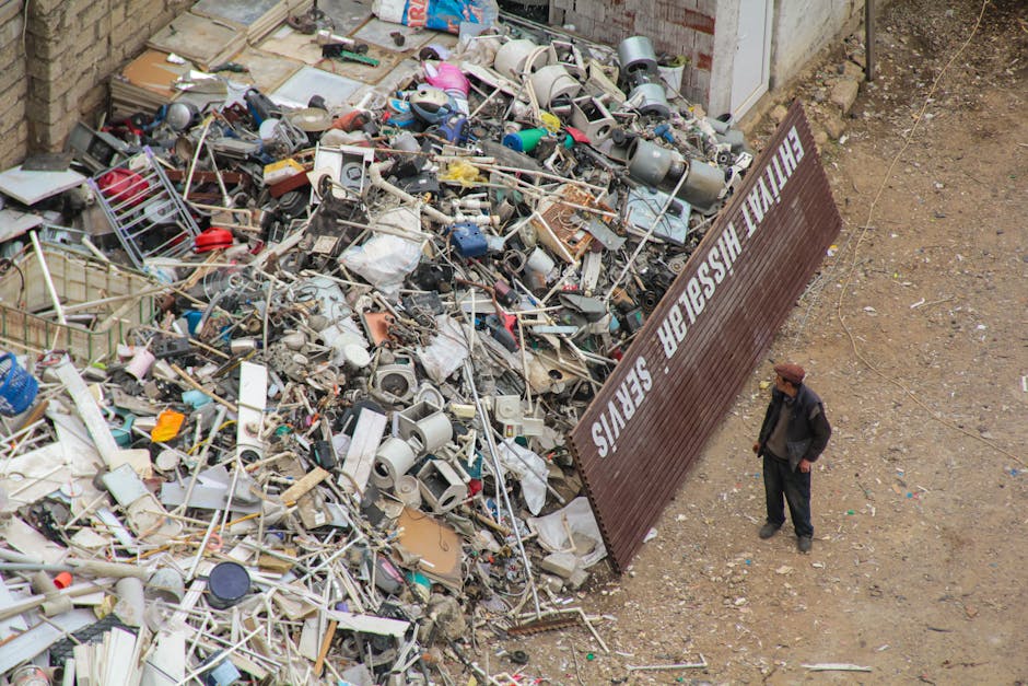 A large green waste disposal container with visible wear and rust on its surface, positioned on a sidewalk. The container is filled with various discarded materials, including flattened cardboard boxes, some with printed labels, and other packaging debris. The cardboard boxes are of different sizes, some open and leaning outward, revealing a mix of plain and printed exteriors in brown and white tones. To the right of the container, there is a wooden pallet stacked with more cardboard, leaning against the side of the container. In the background, blurred elements of an urban environment are visible, including a storefront window, a potted plant, and a parked vehicle on the street. The scene suggests an area undergoing private rubbish collection or on-site clearance, possibly handled by a professional waste removal service such as Rubbish Removal Haringey, with typical materials intended for disposal or recycling.