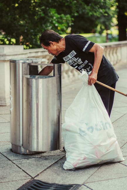 A middle-aged man with short dark hair and a focused expression is seen wearing a black T-shirt with white text and navy trousers, standing outdoors on a paved walkway lined with greenery and trees in the background. He is holding a long wooden stick in his right hand and using his left hand to pick up a large, white, plastic trash bag filled with rubbish, which is partially spilling onto the pavement. The man is leaning forward, actively engaged in rubbish collection or waste management. In front of him are three sleek, stainless steel trash bins arranged side by side, each with a rectangular opening at the top, supported on a concrete base. The environment suggests it might be a park or public space where waste is being managed as part of ad hoc rubbish removal, perhaps as an alternative to scheduled collection services. The overall scene presents a professional yet informal approach to waste disposal and rubbish removal, supported by the presence of the company Rubbish Removal Haringey, which offers services related to rubbish clearance in the area.