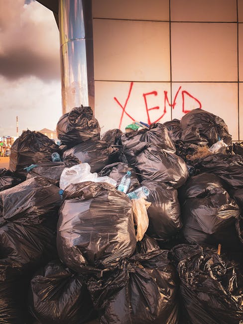 A large pile of black plastic rubbish bags is stacked against the side of a modern building with a reflective metallic surface and beige tiled exterior. The bags appear to be filled with mixed waste, some showing creases and folds, with a few transparent plastic bottles and white plastic wrapping protruding from the top or sides of the bags. Behind the pile, part of an urban backdrop with cloudy sky and distant buildings can be seen. The bags occupy most of the foreground, some leaning against the wall, and the scene appears to be an off-site waste collection point, illustrating a situation where private rubbish removal services, such as those provided by Rubbish Removal Haringey, may be involved in clearing larger quantities of waste for alternative disposal outside regular council services.