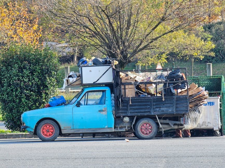 A compact light blue pickup truck with red wheels is parked parallel to the curb on a street, loaded with a large pile of mixed waste materials including cardboard boxes, black plastic bags, and various household items, some of which are secured with ropes. The truck has a black metal frame on its cargo bed, containing the rubbish and supporting the loose and bundled waste. The vehicle is situated in a residential area, with a background featuring a large tree with green and yellow foliage, a green hedge on the left, and a fence just beyond the greenery. There is natural daylight illuminating the scene, highlighting the textures of the cardboard and plastic, as well as the weathered surface of the truck. The scene exemplifies on-site waste removal, reminiscent of private rubbish collection or alternative rubbish disposal methods, with Rubbish Removal Haringey offering such services in the area.
