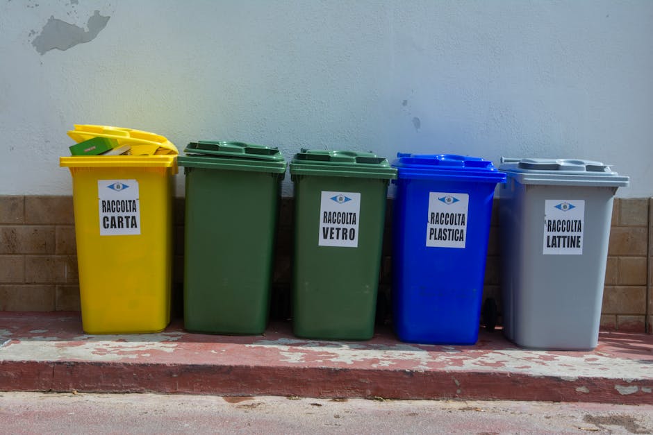 A row of five colour-coded recycling bins positioned on a paved sidewalk in front of a light grey wall and a brick base. From left to right, the first bin is yellow with a closed lid, labeled for paper collection ('RACCOLTA CARTA'), with some yellow recyclables visible on top. Next are two green bins without visible lids, designated for glass ('RACCOLTA VETRO'), placed side by side. The fourth bin is blue with a closed lid, labeled for plastic waste ('RACCOLTA PLASTICA'), and the last bin is grey with a closed lid, intended for containers like cans ('RACCOLTA LATTINE'). The bins are made of sturdy plastic with a textured finish, and they rest directly on the sidewalk surface, which shows signs of weathering and slight paint peeling near the curb. The scene appears to be outdoors during daytime, providing a clear view of how these bins would be used for on-site waste separation, aligning with private waste disposal practices apart from local authority collection methods. Rubbish Removal Haringey's service practices in waste segregation photography are subtly reflected here through the organized display of waste containers for efficient rubbish management.