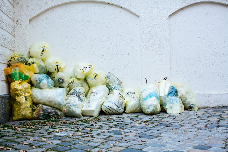 A collection of various garbage bags filled with waste materials, arranged in a neat pile against a white exterior wall on a cobbled pavement. The bags are made of translucent plastic, showing glimpses of their contents, and come in different colours including yellow, white, and clear, with some tied securely with black or yellow plastic ties. The bags vary in size and shape, with some appearing full and tightly packed, while others are slightly less firm. The cobblestone ground beneath the bags consists of small, uneven stones in shades of grey and brown, with some fallen leaves scattered nearby. The background features a smooth, white painted wall with subtle paneling or architectural recesses, and a corner of a brick wall or building is visible on the left side. The scene is evenly lit, suggesting natural daylight, illustrating waste awaiting collection or disposal, relevant to private rubbish removal services such as those offered by Rubbish Removal Haringey, highlighting the need for efficient waste management solutions.